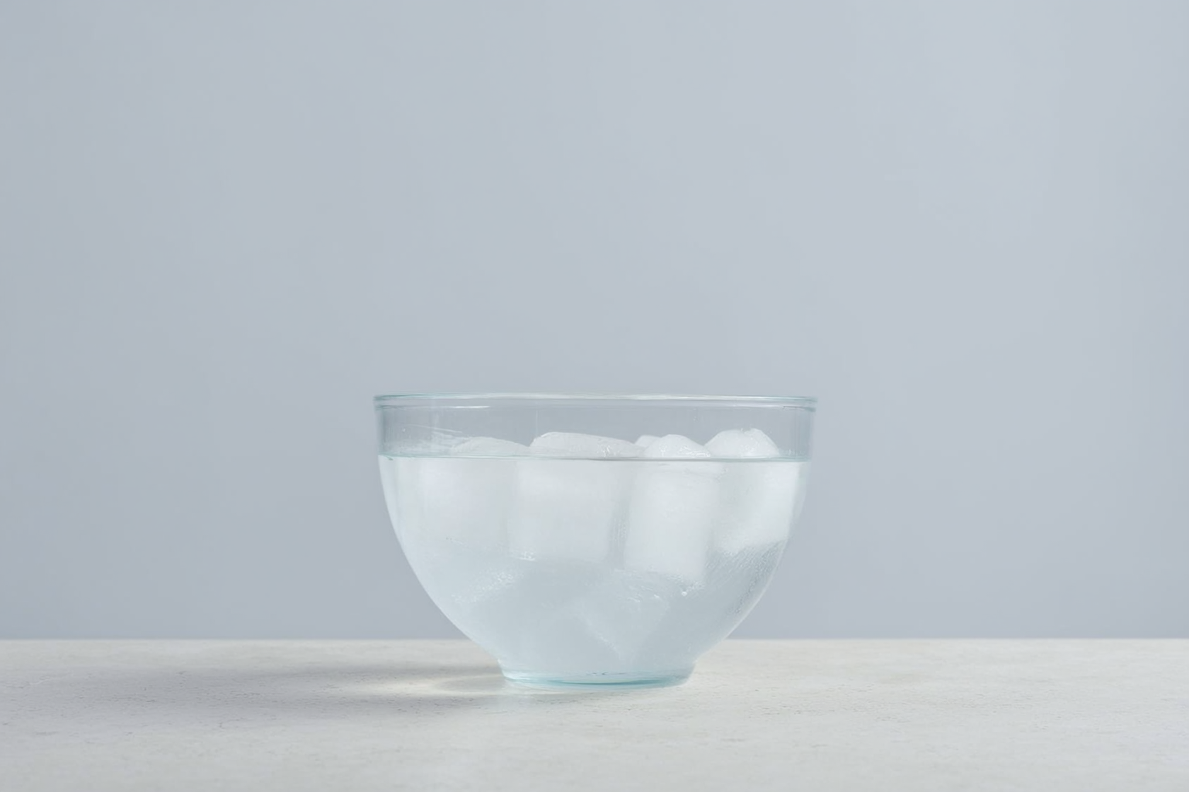 Eine klare Glasschüssel mit Eiswasser auf einer hellen Steinoberfläche vor ruhigem, einfarbigem Hintergrund.